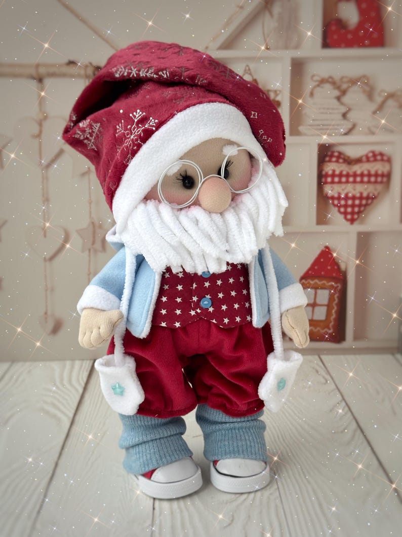 May include: A handmade Santa Claus doll wearing a red hat with snowflake embroidery, a white beard, and round glasses. He has a red vest with white stars, red pants, a light blue jacket, and white mittens. The doll is wearing white shoes with red soles.