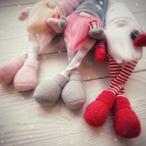 Pu&ograve; includere: Tre gnomi di peluche con cappelli rosa, bianchi e grigi e abiti abbinati. Gli gnomi sono seduti su una superficie di legno bianco con uno sfondo scintillante.