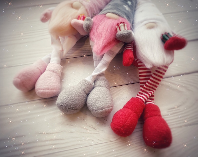 Pu&ograve; includere: Tre gnomi di peluche con cappelli rosa, bianchi e grigi e abiti abbinati. Gli gnomi sono seduti su una superficie di legno bianco con uno sfondo scintillante.