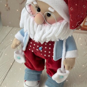 May include: A handmade Santa Claus doll wearing a red hat and pants with white star patterns, a blue jacket, and white mittens. The doll has a white beard, glasses, and blue sneakers. A festive Christmas decoration.