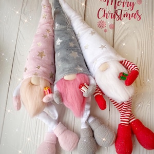 Pu&ograve; includere: Tre gnomi di Natale con gambe lunghe e cappelli a punta. Gli gnomi indossano cappelli rosa, grigi e bianchi con stelle dorate. Gli gnomi tengono piccole scatole regalo rosse. Gli gnomi sono seduti su una superficie di legno bianco. Il testo "Merry Christmas" &egrave; scritto in corsivo rosso sopra gli gnomi.