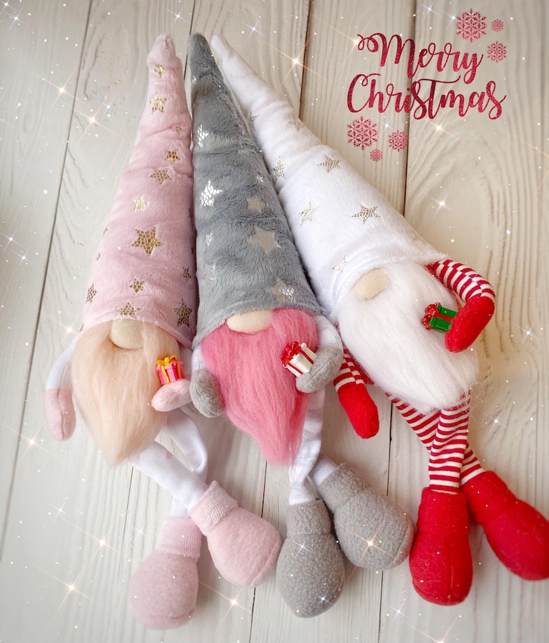 Pu&ograve; includere: Tre gnomi di Natale con gambe lunghe e cappelli a punta. Gli gnomi indossano cappelli rosa, grigi e bianchi con stelle dorate. Gli gnomi tengono piccole scatole regalo rosse. Gli gnomi sono seduti su una superficie di legno bianco. Il testo "Merry Christmas" &egrave; scritto in corsivo rosso sopra gli gnomi.