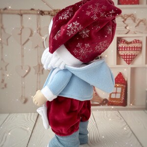 May include: A handmade doll dressed in a festive outfit. The doll wears a red velvet hat with silver snowflake embroidery, a light blue jacket, red velvet pants, and blue leg warmers. The doll is standing on a wooden surface.