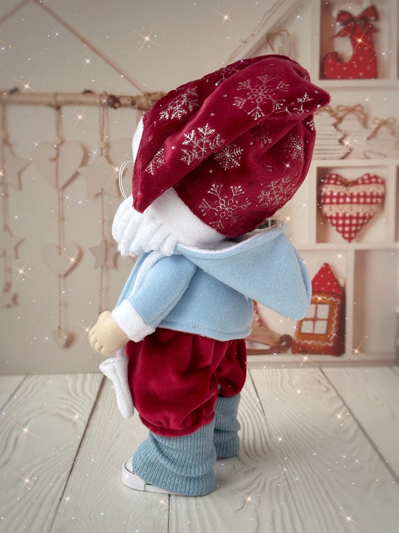 May include: A handmade doll dressed in a festive outfit. The doll wears a red velvet hat with silver snowflake embroidery, a light blue jacket, red velvet pants, and blue leg warmers. The doll is standing on a wooden surface.