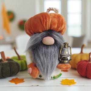 May include: A plush gnome doll with a pumpkin hat and a long gray beard. The gnome holds a miniature lantern and has a round nose. The gnome is surrounded by other decorative pumpkins in various colors, including green, orange, and red.