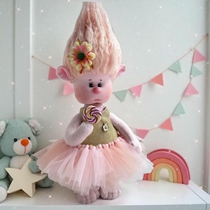 May include: A handmade doll with a pink tulle skirt, holding a swirl lollipop. The doll has a large pink hair, a flower, and a gold vest. A plush bear and a rainbow decoration are in the background.