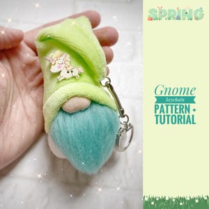 May include: A handmade gnome keychain with a light green plush hat and a blue beard. The gnome has a wooden unicorn button attached to the hat. The keychain also has a silver-colored metal clasp. The image includes the text "Spring" and "Gnome keychain PATTERN + TUTORIAL".
