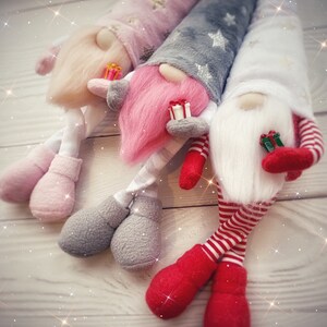 Pu&ograve; includere: Tre gnomi di peluche con lunghe barbe bianche e cappelli rossi, rosa e grigi. Gli gnomi tengono piccole scatole regalo e sono seduti su una superficie di legno bianco.