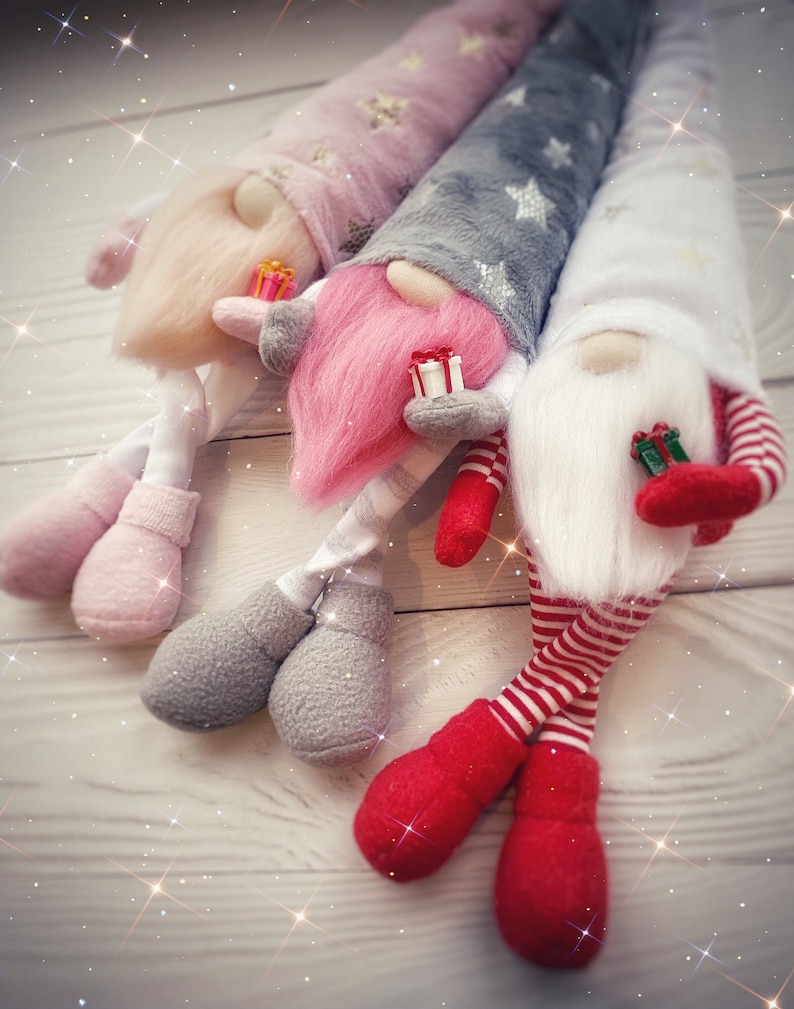 Pu&ograve; includere: Tre gnomi di peluche con lunghe barbe bianche e cappelli rossi, rosa e grigi. Gli gnomi tengono piccole scatole regalo e sono seduti su una superficie di legno bianco.