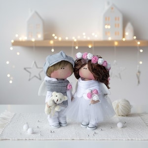 Może przedstawiać: Dwie ręcznie robione lalki z tkaniny, chłopiec i dziewczynka, stoją obok siebie. Chłopiec ma na sobie szarą czapkę i szalik, trzymając misia. Dziewczynka ma na sobie białą sukienkę i wianek z kwiatów, trzymając bukiet.