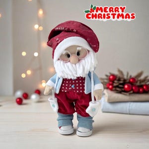 May include: A handmade Santa Claus doll wearing a burgundy hat, red pants, and a blue jacket. The doll has a white beard, glasses, and mittens. The text "MERRY CHRISTMAS" is above the doll.