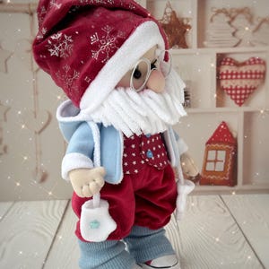 May include: A handmade Santa Claus doll wearing a red velvet hat with silver snowflake accents, a white beard, and round glasses. He is dressed in a blue jacket, red pants, and blue leg warmers, holding a white mitten with a blue star. The doll is wearing white sneakers.