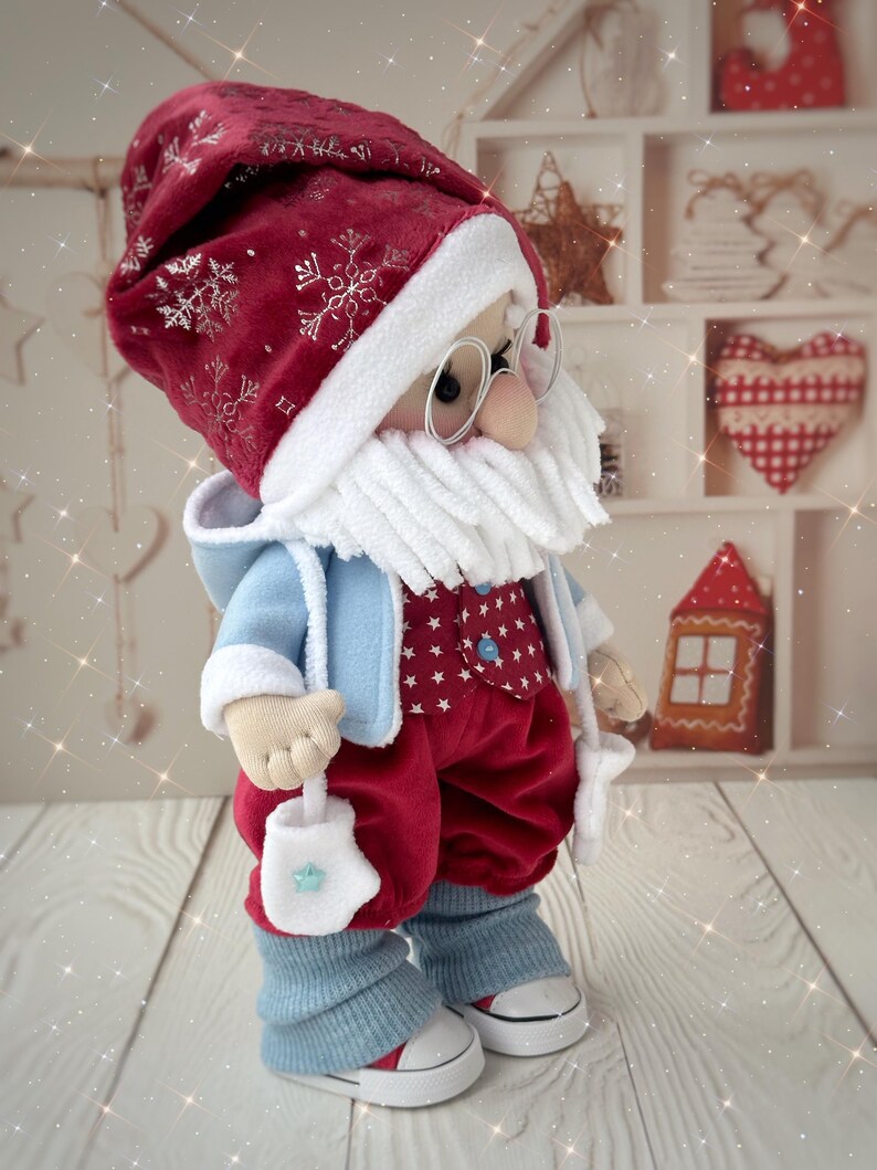 May include: A handmade Santa Claus doll wearing a red velvet hat with silver snowflake accents, a white beard, and round glasses. He is dressed in a blue jacket, red pants, and blue leg warmers, holding a white mitten with a blue star. The doll is wearing white sneakers.