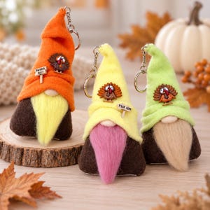 May include: Three gnome keychains with felt hats in orange, yellow, and light green. Each gnome features a turkey embellishment and a long beard. The orange gnome has a sign reading PARDON ME, and the yellow gnome has a sign reading PICK HIM.