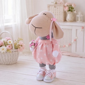 May include: A plush sheep doll with a tan face and floppy ears, wearing a pink dress with a white star pattern, a grey turtleneck collar, and grey leggings. The doll has a small pink heart accessory on its head and wears pink sneakers.