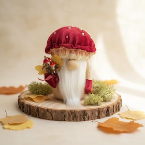 Könnte beinhalten: Eine skurrile Pilz-Wichtel-Figur mit einer roten Samtkappe, die mit Perlen verziert ist. Der Wichtel hält einen Strauß Herbstblätter und Miniaturpilze. Er sitzt auf einer Holzscheibe, umgeben von Moos und fallenden Blättern.
