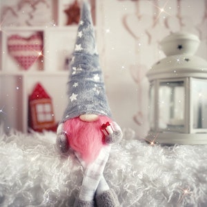 Pu&ograve; includere: Un gnomo grigio e bianco con la barba rosa e una scatola regalo rossa e bianca &egrave; seduto su una superficie bianca e soffice. Il gnomo indossa un cappello grigio con stelle bianche e un abito a righe grigio e bianco. Lo sfondo &egrave; sfocato e presenta uno scaffale di legno bianco con un cuore rosso e bianco e una lanterna bianca.