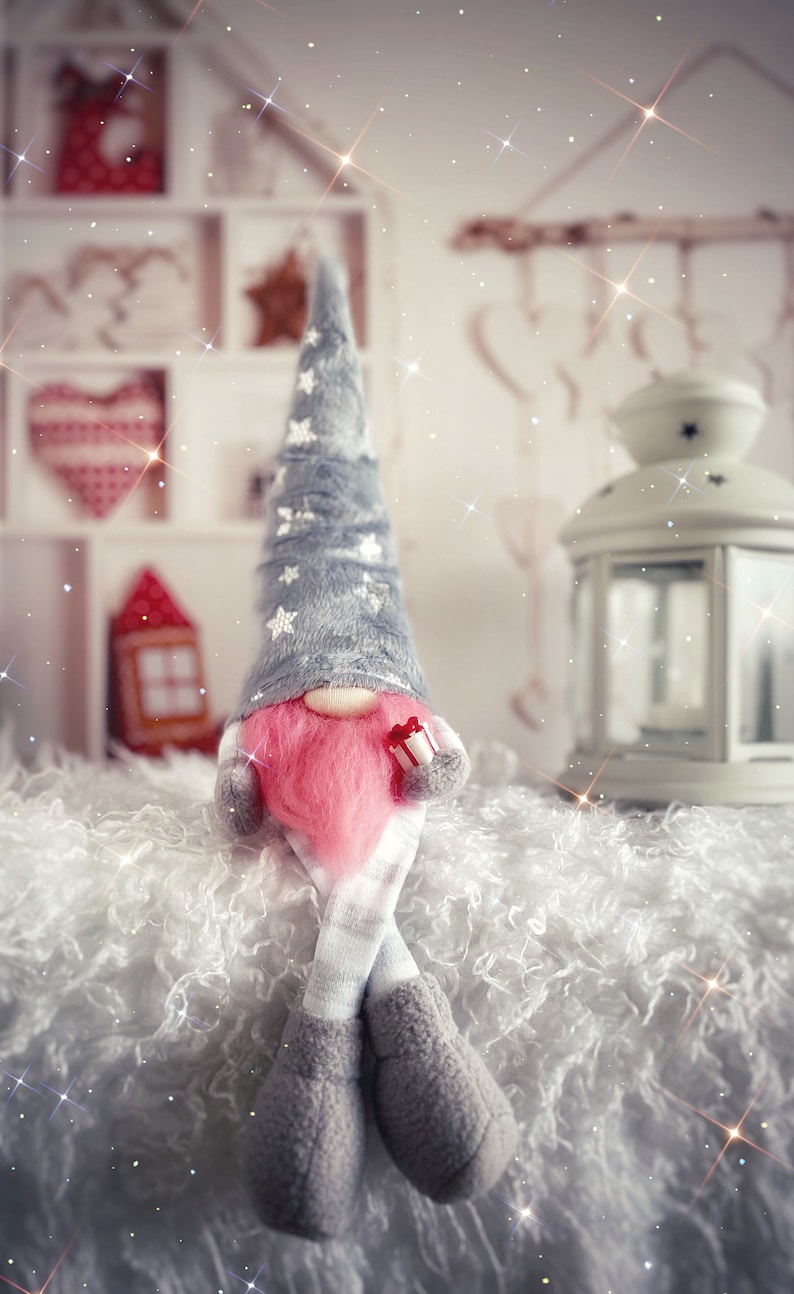 Pu&ograve; includere: Un gnomo grigio e bianco con la barba rosa e una scatola regalo rossa e bianca &egrave; seduto su una superficie bianca e soffice. Il gnomo indossa un cappello grigio con stelle bianche e un abito a righe grigio e bianco. Lo sfondo &egrave; sfocato e presenta uno scaffale di legno bianco con un cuore rosso e bianco e una lanterna bianca.