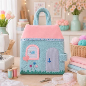 Könnte beinhalten: Eine hellblaue und rosafarbene Stofftasche in Hausform mit einer texturierten Fleece-Außenseite. Die Tasche verfügt über ein rosafarbenes Dach, ein Fenster mit Wal-Grafik, eine graue Tür mit blauem Anker, einen herzförmigen Ballon und Blumen.