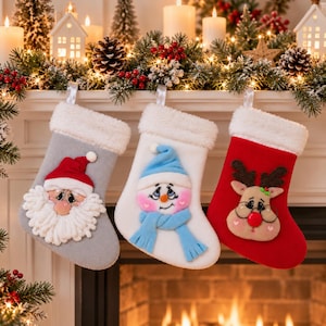 Op de afbeelding: Drie kerstsokken met witte randen van imitatiebont. De grijze sok heeft een kerstman-gezicht, de witte sok toont een sneeuwpop met een blauwe muts en sjaal, en de rode sok toont een rendier-gezicht met een bril en een rode neus.