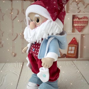 May include: A handmade Santa Claus doll, featuring a red hat with snowflake accents, a white beard, and round glasses. The doll wears a blue coat, red pants with white stars, and blue leg warmers over white sneakers. A festive Christmas decoration.