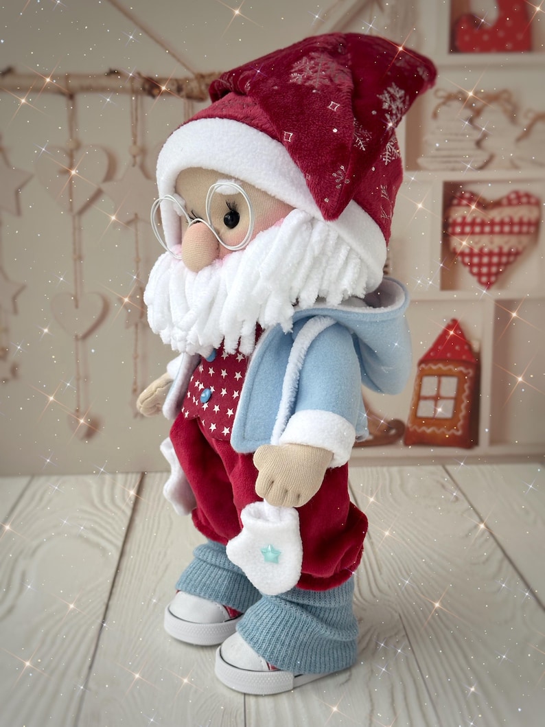 May include: A handmade Santa Claus doll, featuring a red hat with snowflake accents, a white beard, and round glasses. The doll wears a blue coat, red pants with white stars, and blue leg warmers over white sneakers. A festive Christmas decoration.