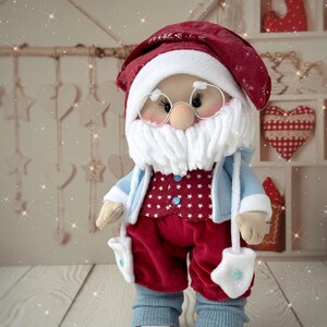 May include: A handmade Santa Claus doll with a red hat and pants, a white beard, and round glasses. The doll wears a blue jacket over a red star-patterned vest, with blue leg warmers and white sneakers. The doll has white mittens.