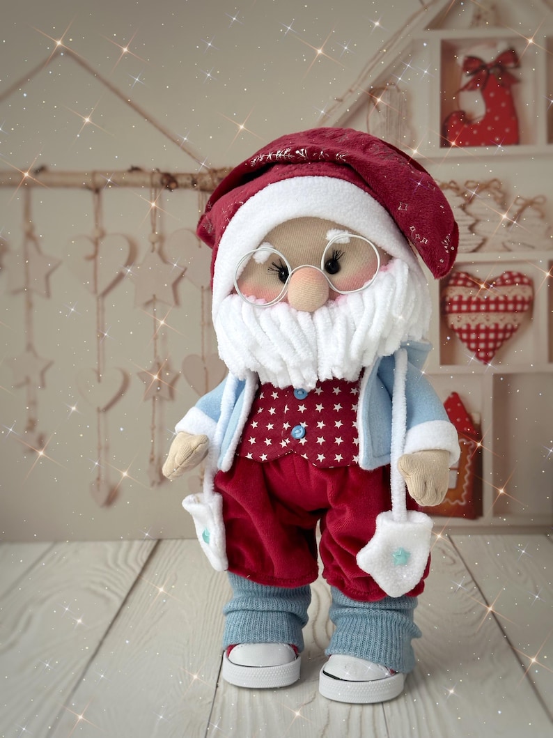 May include: A handmade Santa Claus doll with a red hat and pants, a white beard, and round glasses. The doll wears a blue jacket over a red star-patterned vest, with blue leg warmers and white sneakers. The doll has white mittens.