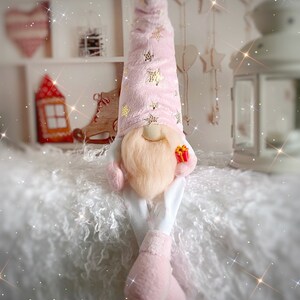 Pu&ograve; includere: Un gnomo rosa con la barba bianca e un cappello rosa con stelle dorate &egrave; seduto su una superficie bianca e soffice. Lo gnomo tiene una piccola scatola regalo rossa.
