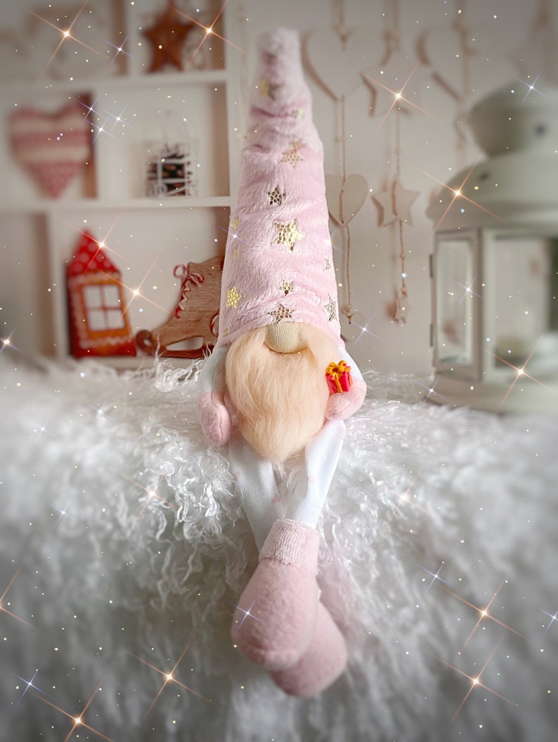 Pu&ograve; includere: Un gnomo rosa con la barba bianca e un cappello rosa con stelle dorate &egrave; seduto su una superficie bianca e soffice. Lo gnomo tiene una piccola scatola regalo rossa.