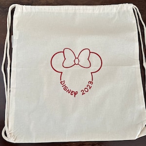 Puede incluir: Una bolsa de cordón blanco con un diseño bordado en rojo de la cabeza de Mickey Mouse con un lazo y el texto "DISNEY 2023".