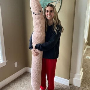 Peut inclure: Une grande peluche en forme de ver, rose clair, crochetée, avec un visage simple. La peluche est maintenue debout. Le jouet mesure environ 152 cm de haut et est fait d'un fil doux et texturé.