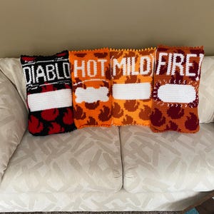 Puede incluir: Cuatro cojines de ganchillo con forma de bolsas de patatas fritas, cada uno con una etiqueta de sabor diferente: "DIABLO", "HOT", "MILD" y "FIRE". Los cojines presentan un rectángulo blanco para el nombre de la patata y un diseño estampado en rojo, naranja y marrón.