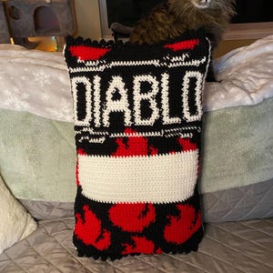 May include: A rectangular crochet pillow with the word "DIABLO" in white block letters on a black background. The pillow features red heart-shaped designs and a white stripe. The pillow is on a bed with a cat.