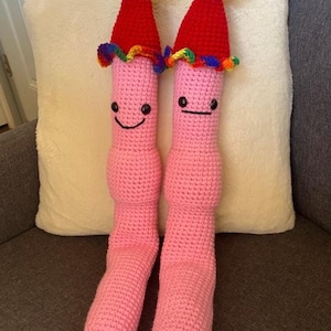 May include: Two handmade pink crochet worm-shaped dolls. Each has a red party hat with a rainbow pom-pom and a rainbow trim. The worms have black eyes and a smiling mouth. The dolls are sitting on a gray couch with a white pillow.