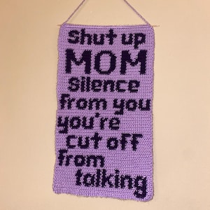 以下が含まれることがあります： 手作りのラベンダー色の編み物ウォールハンギング。テキストは「shut up MOM silence from you you're cut off from talking」と濃い紫色の文字で書かれています。薄紫色の紐で吊るされています。