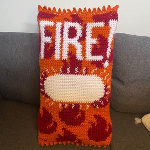 Könnte beinhalten: Gehäkeltes Kissen in Form einer Snack-Tüte mit dem Wort "FIRE!" in Weiß. Das Kissen hat einen orangefarbenen Hintergrund mit roten Flammenmustern und eine weiße ovale Form. Die Kanten sind gewellt.