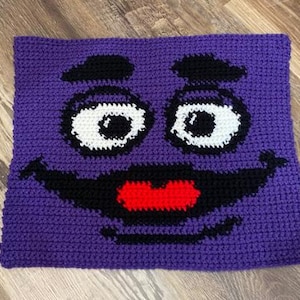 May include: A handmade crochet pillow featuring a smiling face design. The pillow has a purple background with black eyebrows, a black mouth, white eyes, and a red mouth. The pillow is square-shaped and appears to be made of yarn.