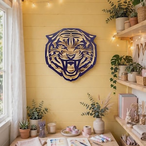 Puede incluir: Una pieza de arte mural de madera en capas que presenta una cabeza de tigre estilizada con expresión rugiente. El diseño consta de un marco exterior de color morado oscuro y una capa interior de madera natural con recortes detallados.