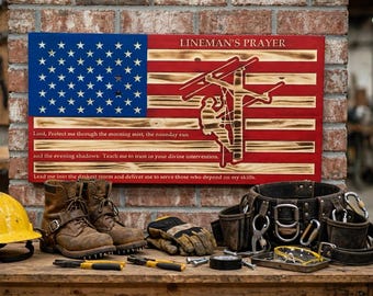 May include: A rectangular wooden sign featuring a painted American flag, a silhouette of a lineman on a utility pole, and the text LINEMAN'S PRAYER with a prayer inscribed below. The sign is displayed above a yellow hard hat, brown work boots, gloves, and a leather tool belt.
