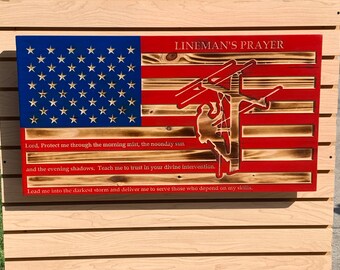 May include: A wooden American flag with a red, white, and blue color scheme. The flag has a silhouette of a lineman working on a power line. The text "LINEMAN'S PRAYER" is at the top of the flag. The text below the silhouette reads: "Lord, Protect me through the morning mist, the noonday sun and the evening shadows. Teach me to trust in your divine intervention. Lead me into the darkest storm and deliver me to serve those who depend on my skills."