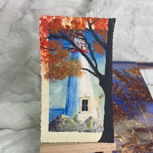 Könnte beinhalten: Ein kleines rechteckiges Aquarellbild auf strukturiertem Papier, das einen weißen Leuchtturm mit blauen Akzenten vor einem Hintergrund aus orangefarbenem Herbstlaub und einer dunklen Baumsilhouette zeigt, auf einer Miniatur-Holzstaffelei.