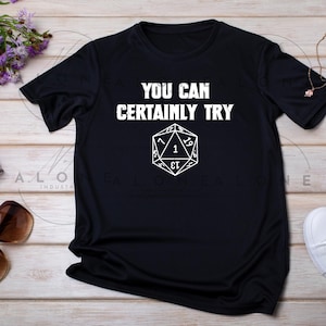 Puede incluir: Camiseta negra con texto blanco "YOU CAN CERTAINLY TRY" sobre un gráfico de dado D20 blanco. El gráfico del dado tiene el número 1 en el centro. La camiseta se muestra sobre una superficie clara de vetas de madera.