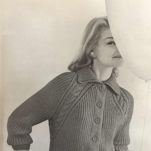 May include: A close-up of a woman wearing a gray knitted cardigan with a cable knit pattern and button closure. The cardigan has a collared neckline and long sleeves.