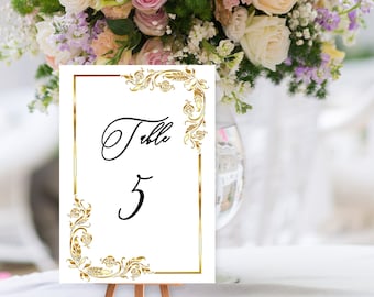 Könnte beinhalten: Eine weiße Tischnummerkarte mit goldenen Filigranecken und einem goldenen Rahmen. Das Wort "Table" ist in eleganter schwarzer Schrift über der Zahl "5" geschrieben. Die Karte wird auf einer kleinen Holzstaffelei mit einem verschwommenen Blumenarrangement im Hintergrund angezeigt.