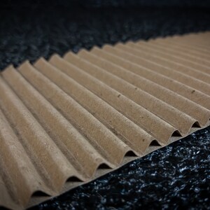 May include: A close-up of a brown corrugated cardboard sheet with a wavy texture. The sheet is lying on a black surface.