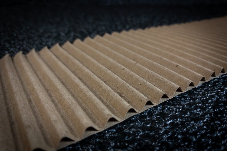 May include: A close-up of a brown corrugated cardboard sheet with a wavy texture. The sheet is lying on a black surface.