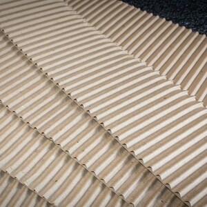 May include: Close-up of corrugated cardboard sheets, showing the texture and pattern of the material. The sheets are stacked in a diagonal pattern, creating a visual effect of depth and dimension.