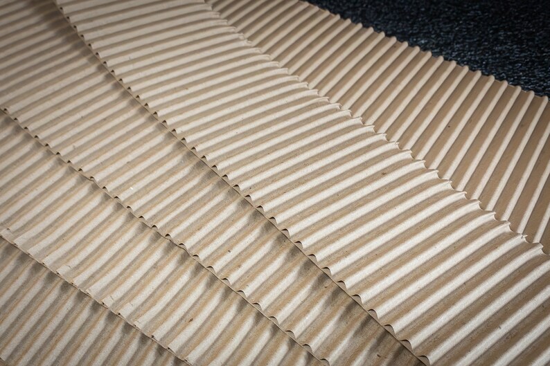 May include: Close-up of corrugated cardboard sheets, showing the texture and pattern of the material. The sheets are stacked in a diagonal pattern, creating a visual effect of depth and dimension.