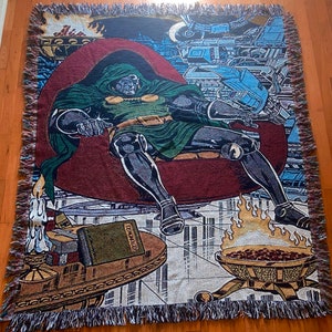 Op de afbeelding: Een geweven wandkleed met een superheldenfiguur, Doctor Doom, zittend op een troon met een boek voor hem. Het wandkleed is in tinten groen, rood en bruin met een gedetailleerde achtergrond.